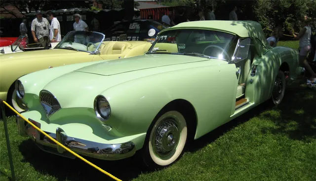 Front three-quarter view of a Pine Tint Darrin with the Deauville top raised, showing the production windshield, top profile, pocket door, and roadster proportions.