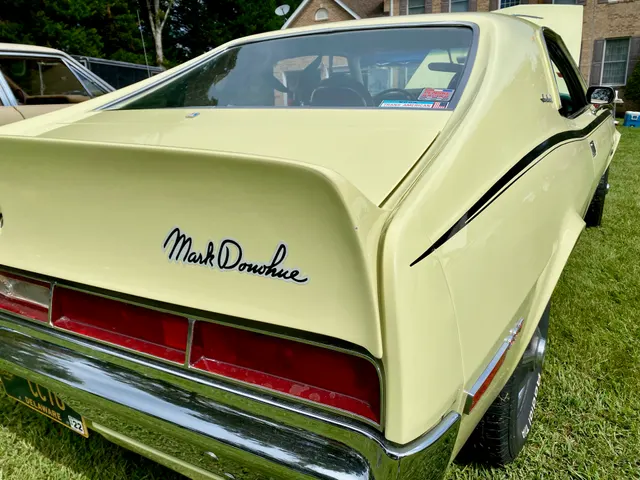 Same-car close-up of the yellow paint, black side stripe, ducktail spoiler, Mark Donohue signature, rear lamps, and brightwork.
