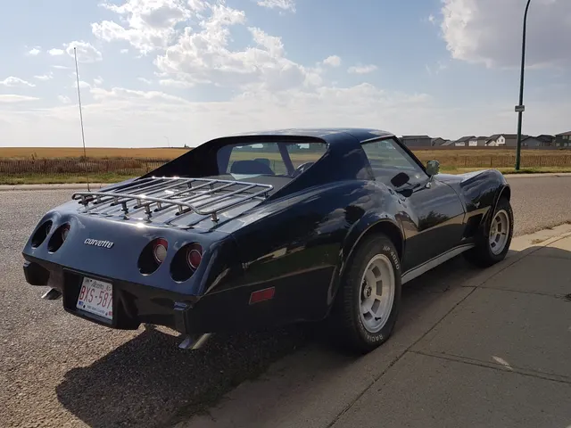 Blue 1977 coupe in rear three-quarter view with a luggage rack and cast-aluminum-style wheels; the photograph shows installed equipment, while records and identifiers are still required to establish factory V54 or YJ8 equipment.