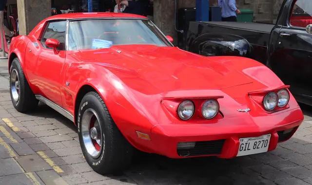 Red 1977 coupe with its concealed headlamps raised, showing the lamp buckets, nose openings, hood, fender vents, roofline, and front wheel treatment; displayed Stingray script and other equipment remain specific to this car.